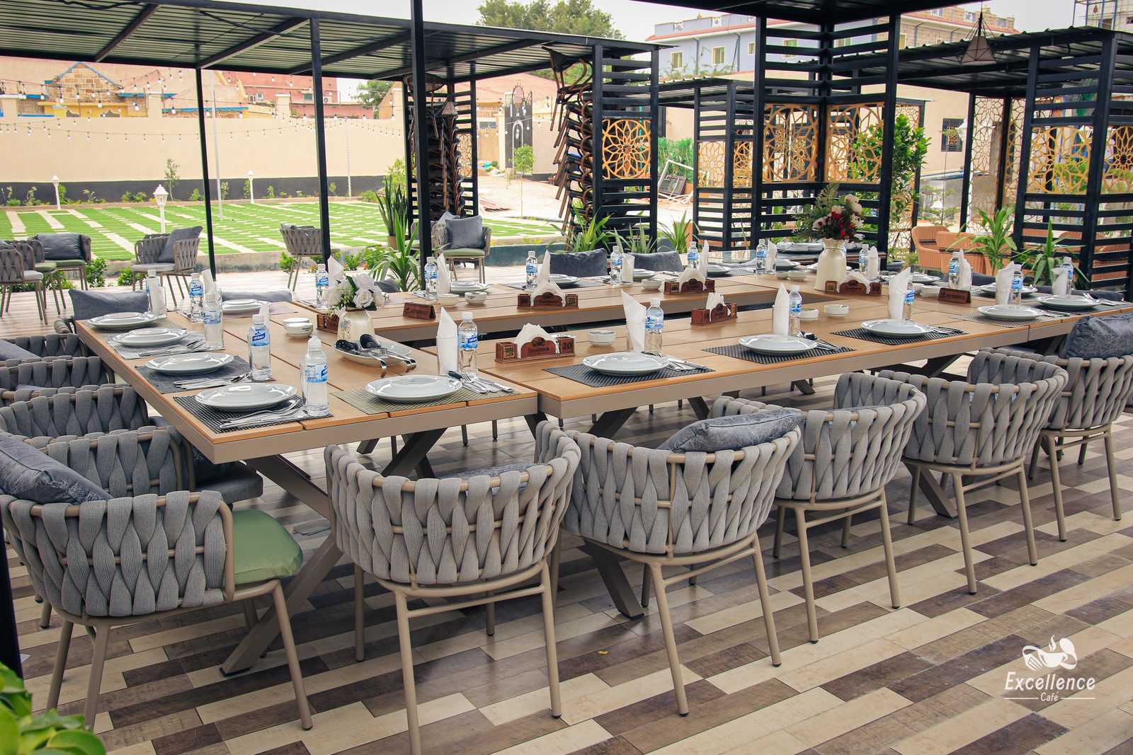 Excellence Café's covered garden terrace — long wooden tables and grey woven armchairs under a black pergola frame, with string lights and a lawn beyond.