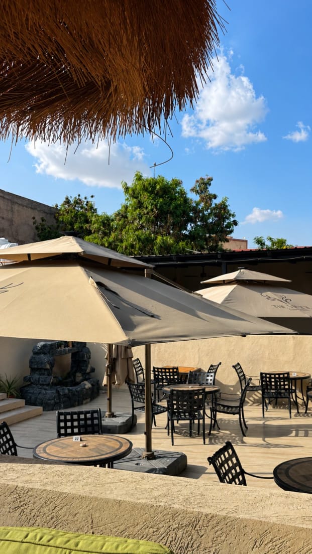 The Village Hargeisa outdoor terrace by day — iron bistro tables shaded by cream parasols, a small stone water feature and a young tree against a blue sky.
