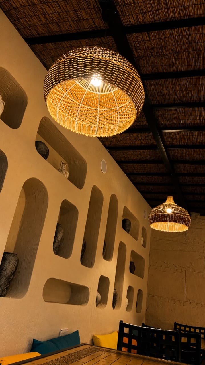 The Village Hargeisa interior feature wall — hand-sculpted adobe with rounded recesses, each holding a smooth stone, beneath woven pendant lamps and a reed ceiling.